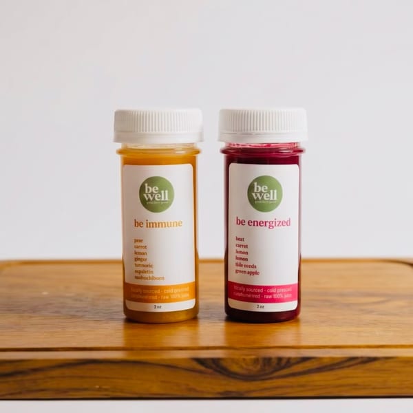 Two Be Well juice shots, "be immune" (orange) and "be energized" (red), on a wooden board.