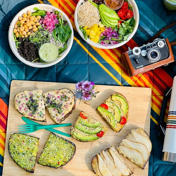 Overhead view of two Be Well Cafe bowls, various toasts, and a vintage camera on a colorful blanket.