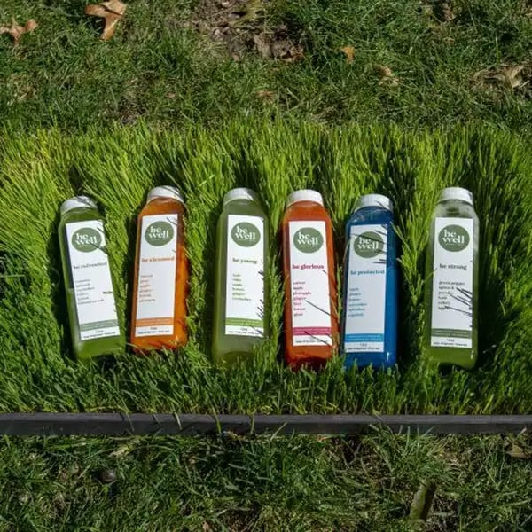 Six colorful Be Well Cafe juice bottles, including 'be refreshed' and 'be strong,' arranged on vibrant green grass.